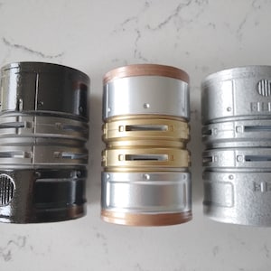 May include: Three cylindrical metal containers with different finishes. The container on the left is black, the middle container is silver with gold accents, and the container on the right is silver.