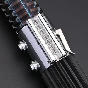 Puede incluir: Primer plano de una empu&ntilde;adura de sable de luz. La empu&ntilde;adura es principalmente negra con detalles plateados y bronce. Cuenta con una serie de peque&ntilde;as luces transparentes y un clip plateado. La parte superior tiene un dise&ntilde;o geom&eacute;trico texturizado.