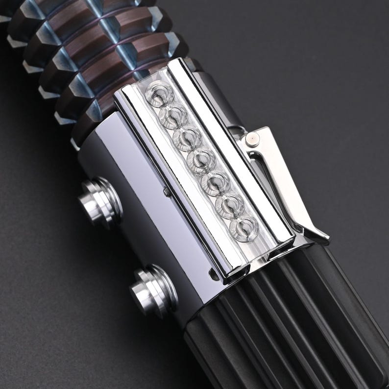 Puede incluir: Primer plano de una empu&ntilde;adura de sable de luz. La empu&ntilde;adura es principalmente negra con detalles plateados y bronce. Cuenta con una serie de peque&ntilde;as luces transparentes y un clip plateado. La parte superior tiene un dise&ntilde;o geom&eacute;trico texturizado.