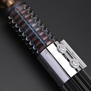 Puede incluir: Primer plano de una empu&ntilde;adura de sable de luz. El agarre marr&oacute;n tiene textura con formas geom&eacute;tricas. El mango tiene una secci&oacute;n plateada con dos botones circulares y las letras "L" y "X". La parte superior es de color bronce.