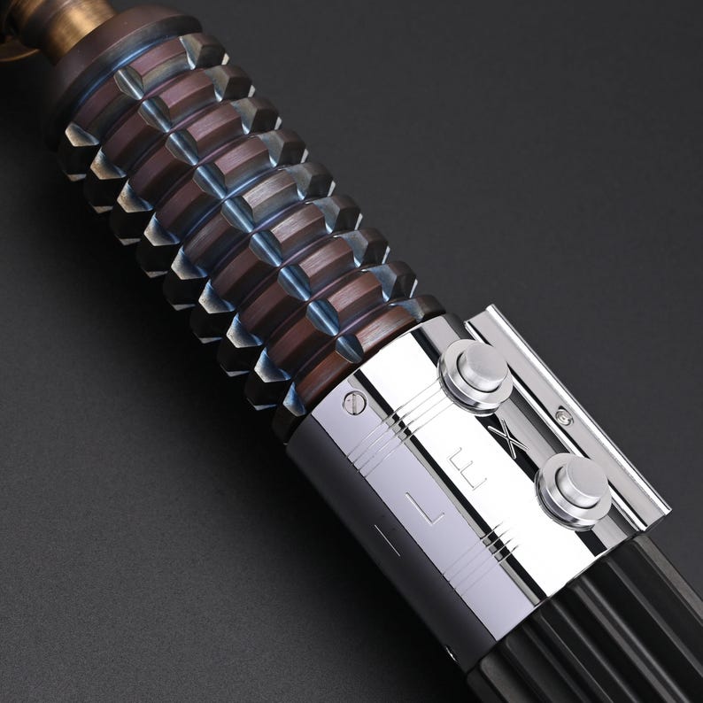 Puede incluir: Primer plano de una empu&ntilde;adura de sable de luz. El agarre marr&oacute;n tiene textura con formas geom&eacute;tricas. El mango tiene una secci&oacute;n plateada con dos botones circulares y las letras "L" y "X". La parte superior es de color bronce.