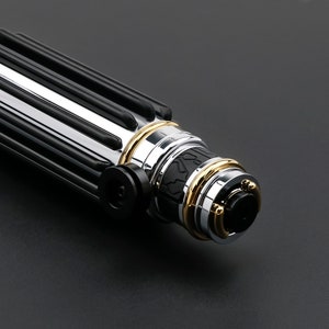 May include: Close-up of a black and silver lightsaber hilt with gold accents. The hilt has a textured grip and a detailed design.