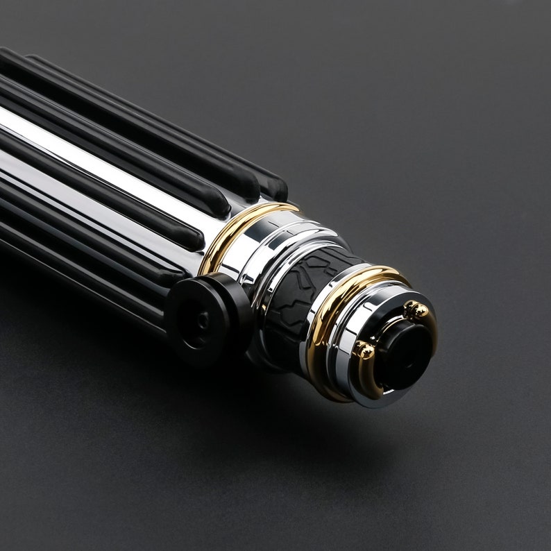 May include: Close-up of a black and silver lightsaber hilt with gold accents. The hilt has a textured grip and a detailed design.