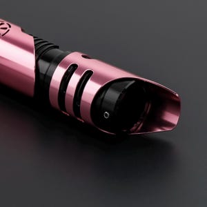 May include: Close-up of a pink and black lightsaber hilt. The hilt has a metallic finish with curved design elements and a black grip section. The lightsaber is set against a dark background.