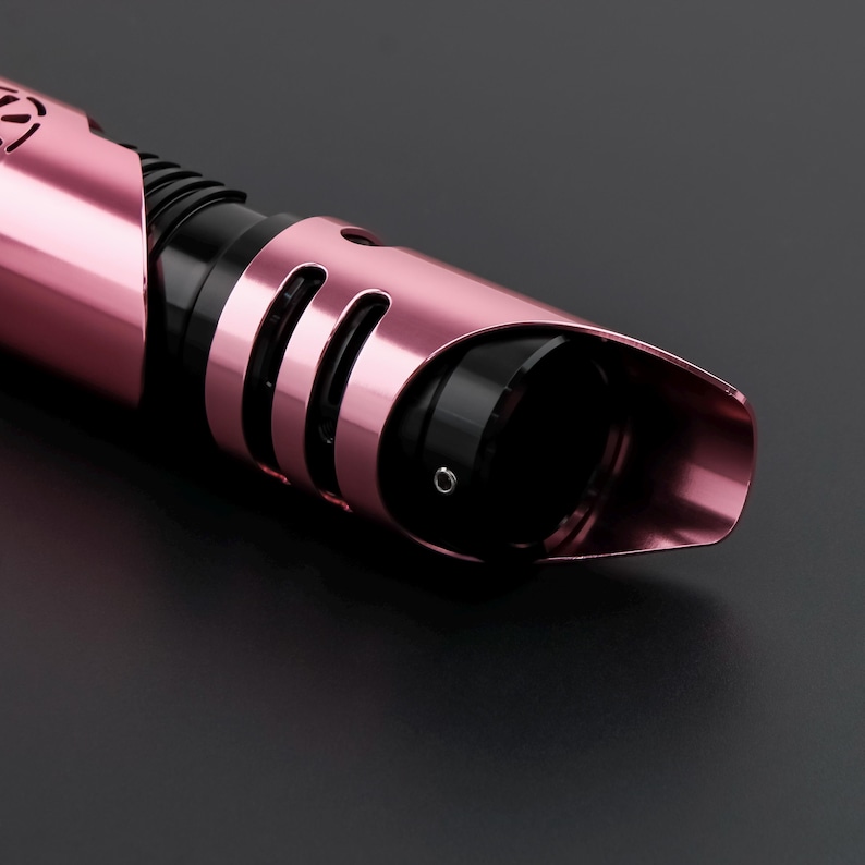 May include: Close-up of a pink and black lightsaber hilt. The hilt has a metallic finish with curved design elements and a black grip section. The lightsaber is set against a dark background.