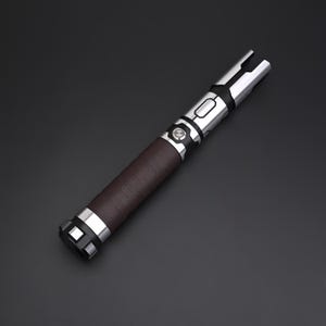 May include: A lightsaber with a brown leather grip, silver accents, and a black button. The blade emitter is silver with a split design. The hilt has a series of silver bands. The lightsaber is a prop or collectible.
