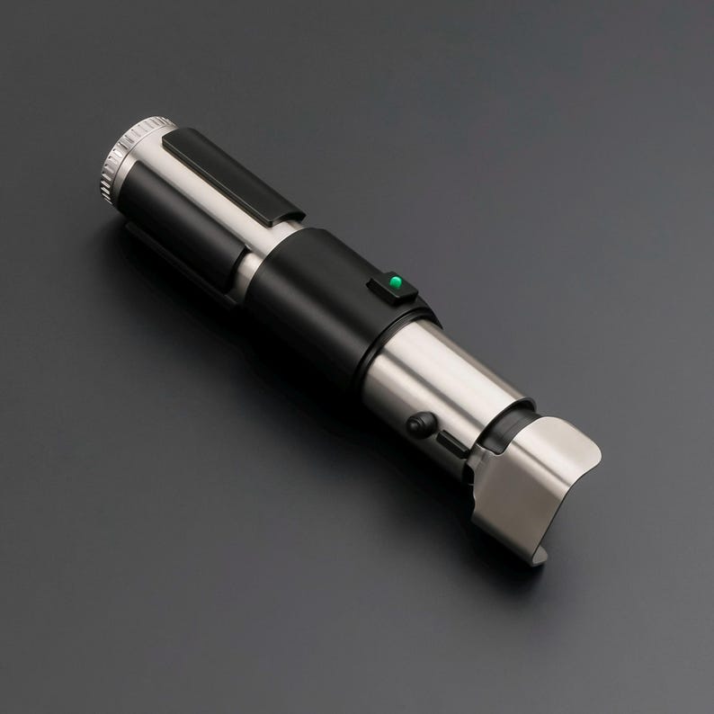 May include: A metallic silver and black lightsaber prop with a green activation button. The prop has a cylindrical design with a curved end and a textured grip. The lightsaber is a replica of a popular science fiction weapon.
