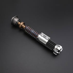 Puede incluir: Un sable de luz met&aacute;lico con componentes plateados, bronce y negros. El mango presenta un dise&ntilde;o acanalado con una secci&oacute;n transparente que revela detalles internos. El sable de luz tiene forma cil&iacute;ndrica y est&aacute; sobre un fondo oscuro.