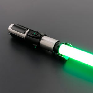 May include: A futuristic lightsaber with a glowing green blade. The hilt is a combination of black, silver, and gray, featuring buttons and a textured grip. The blade emits a bright, vibrant green light, suggesting energy.