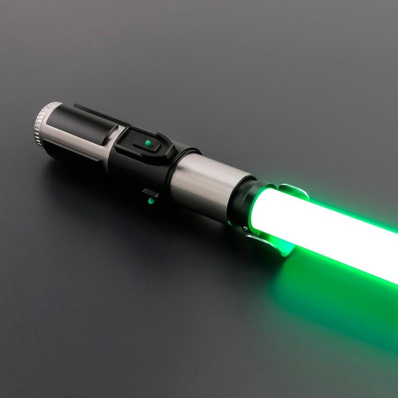 May include: A futuristic lightsaber with a glowing green blade. The hilt is a combination of black, silver, and gray, featuring buttons and a textured grip. The blade emits a bright, vibrant green light, suggesting energy.