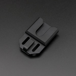 May include: A matte black metal clip with a U-shaped opening and two rectangular slots. The clip has a rectangular shape with a slightly angled top and is designed for secure attachment.