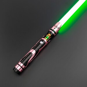 May include: A rose-gold and black lightsaber with a bright green blade. The hilt features intricate detailing, including a black grip section and a gold-colored internal component. The blade emits a vibrant green glow against a dark background.