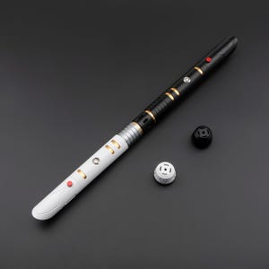 May include: A black and white lightsaber with gold accents and red buttons. The hilt is divided into black and white sections, with gold rings and details. Two detached pieces are next to the lightsaber.