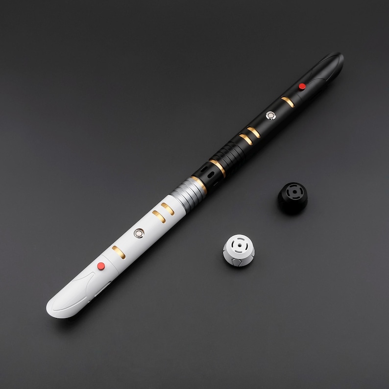 May include: A black and white lightsaber with gold accents and red buttons. The hilt is divided into black and white sections, with gold rings and details. Two detached pieces are next to the lightsaber.
