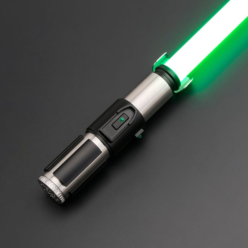 May include: A silver and black lightsaber with a bright green blade. The hilt features a black grip and control panel with a green button. The blade glows brightly, suggesting it is activated. The lightsaber is angled against a dark background.