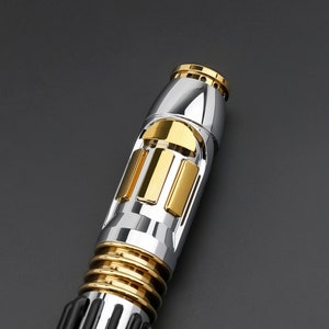 May include: A silver and gold lightsaber replica with a detailed design. The lightsaber has a black handle and a silver blade with gold accents.