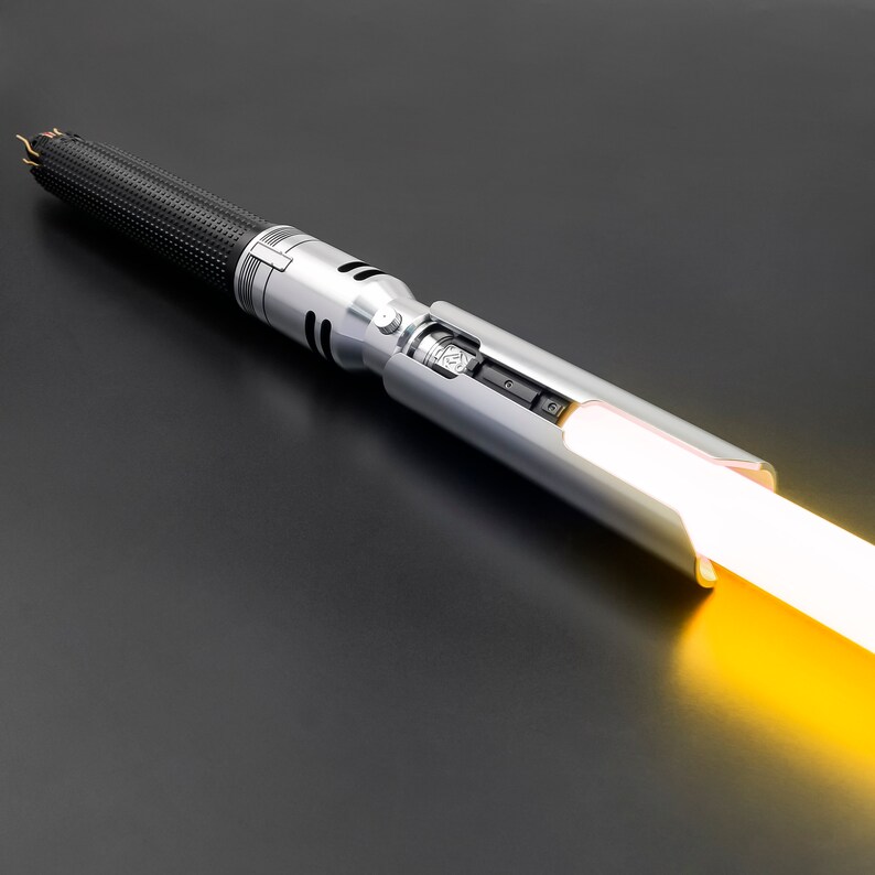 K&ouml;nnte beinhalten: Ein silbernes Lichtschwert mit einem schwarzen Griff und einer leuchtend gelben Klinge. Das Lichtschwert ist eingeschaltet und die Klinge leuchtet.