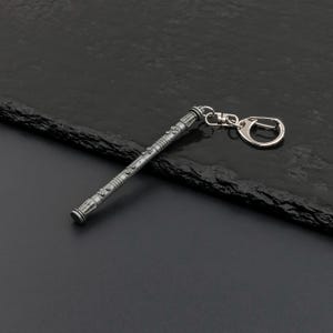 May include: A silver metal keychain with a long, detailed, cylindrical charm attached to a silver split ring.