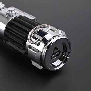 Puede incluir: Primer plano de una empu&ntilde;adura de sable de luz met&aacute;lica. La empu&ntilde;adura presenta una secci&oacute;n negra acanalada, un anillo plateado y una tapa de extremo circular con una letra estilizada. El sable de luz tiene una superficie pulida y reflectante.