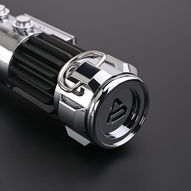 Puede incluir: Primer plano de una empu&ntilde;adura de sable de luz met&aacute;lica. La empu&ntilde;adura presenta una secci&oacute;n negra acanalada, un anillo plateado y una tapa de extremo circular con una letra estilizada. El sable de luz tiene una superficie pulida y reflectante.