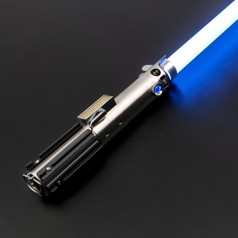 May include: A silver and black lightsaber with a bright blue blade. The hilt features detailed metallic components, including buttons and a rectangular section. The glowing blue blade extends diagonally across a dark background.