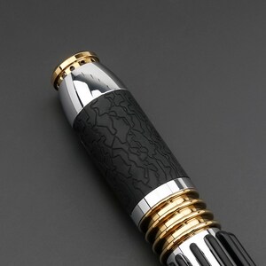 May include: Close-up of a black and silver lightsaber with gold accents. The lightsaber has a textured black grip and a silver blade.