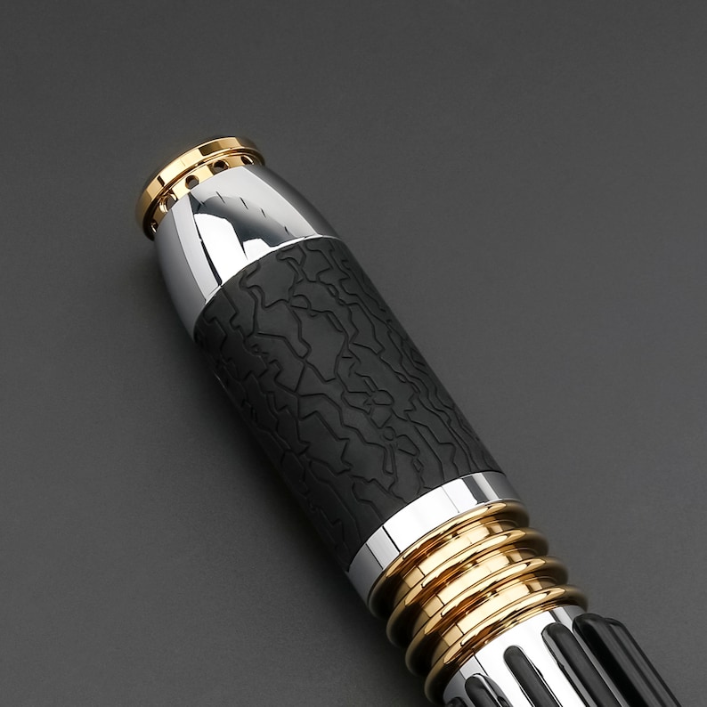 May include: Close-up of a black and silver lightsaber with gold accents. The lightsaber has a textured black grip and a silver blade.