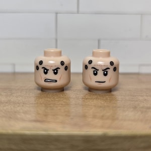 May include: Two tan-colored toy heads with angry expressions. Each head has black markings around the eyes and on the sides, with a furrowed brow and a straight, thin mouth. The heads are on a light brown surface.