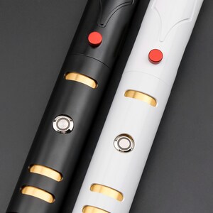 May include: Close-up of two lightsabers, one black and one white. Each has a red button, a silver button, and gold-colored accents. The lightsabers are cylindrical with a sleek, modern design.