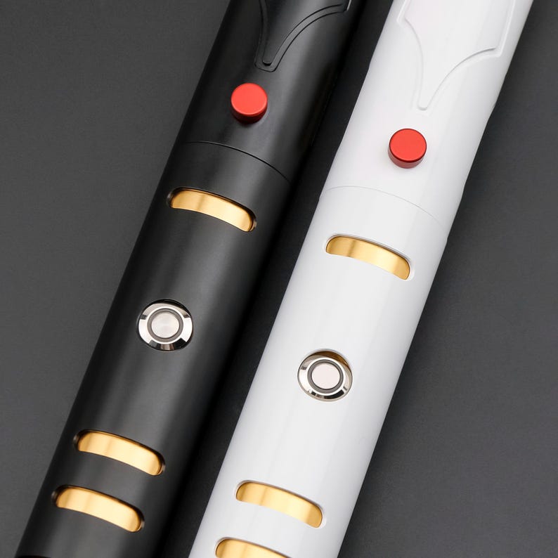May include: Close-up of two lightsabers, one black and one white. Each has a red button, a silver button, and gold-colored accents. The lightsabers are cylindrical with a sleek, modern design.