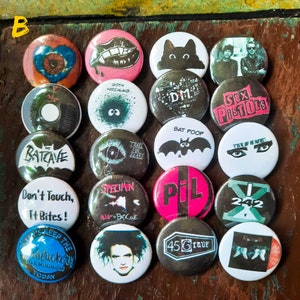 May include: A selection of twenty-one circular pin badges with diverse designs and text. The designs feature a bat, lips, eyes, and band logos. The badges are arranged on a wooden surface.