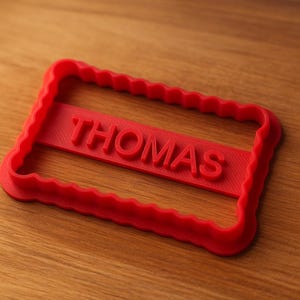 Peut inclure: Emporte-pièce en plastique rouge en forme rectangulaire avec le nom "THOMAS" en relief au centre. Le découpoir a un bord festonné et est posé sur une surface en bois.