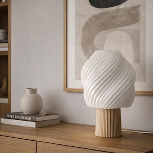 Può includere: Una lampada da tavolo bianca, strutturata, con un design a spirale, poggia su una base in legno. La lampada è su un mobile in legno, con una stampa astratta incorniciata e un piccolo vaso sullo sfondo. Il design della lampada è un elemento di arredo moderno.