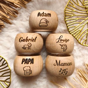 Peut inclure: Cinq ronds de serviette en bois avec des noms et des symboles gravés. Les noms "Adam", "Gabriel", "Louise", "Papa" et "Maman" sont affichés. Chaque anneau a un design unique, dont un bonnet de Père Noël, un lion, une licorne, une chope de bière et des notes de musique.