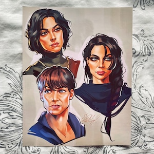 May include: A digital illustration of three women with different hairstyles and clothing. The woman on the left has short, dark hair and is wearing a dark green shirt with a gold chain necklace. The woman in the middle has long, dark hair and is wearing a dark blue shirt. The woman on the right has short, brown hair and is wearing a blue shirt. The illustration is signed "S.A.Y. VERADIA".
