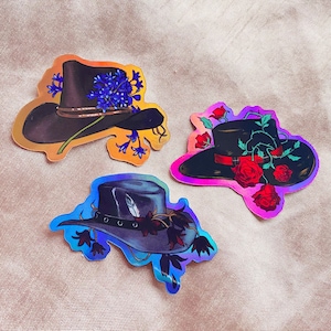 Peut inclure: Trois autocollants holographiques colorés représentant différents chapeaux de cow-boy avec des embellissements floraux. Le premier chapeau est orné de fleurs violettes, le second de roses rouges et le troisième de fleurs violettes foncées.