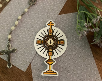 May include: A gold-colored religious icon with a cross and rays of light, on a white background. The icon is a depiction of the monstrance, a sacred vessel used in Catholic worship.