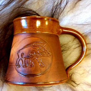 May include: A brown ceramic mug with a raised design of a boar on the front. The mug has a handle and is glazed in a brown colour.