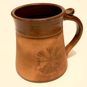 May include: A brown ceramic mug with a brown glaze and a handle. The mug features a compass design with runes.