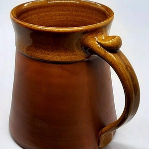 May include: A brown ceramic mug with a wide base and a rounded handle. The mug has a brown glaze with a darker brown rim and handle.