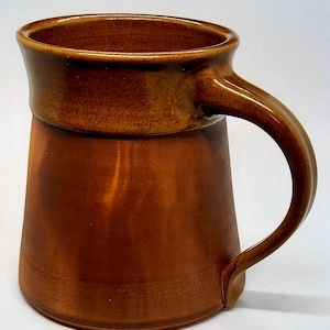 May include: A brown ceramic mug with a wide, rounded base and a slightly flared rim. The mug has a large handle and a speckled glaze.