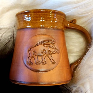 May include: A brown ceramic mug with a raised design of a boar in a circle. The mug has a brown glaze and a handle.