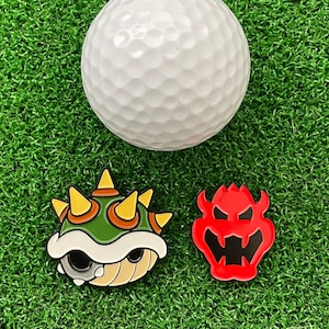May include: A white golf ball positioned above two enamel pins. One pin is a green and yellow spiked shell, and the other pin is a red and black character face with horns. The items are arranged on a textured green surface.