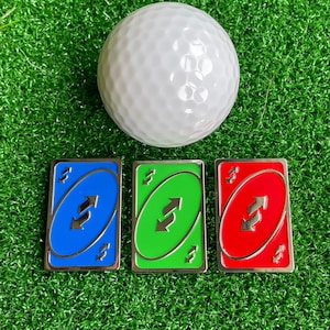 May include: Three Uno Reverse cards, one blue, one green, and one red, are arranged in a row on a green surface. A white golf ball is in the background.