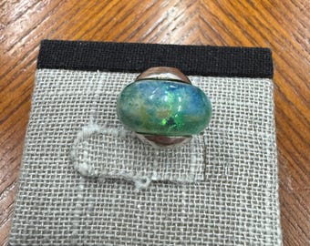 Puede incluir: Un anillo artesanal con una colorida cuenta de vidrio veteado. La cuenta presenta tonos de azul y verde, engastada sobre una base de plata y cobre. El anillo se exhibe sobre una superficie de lino gris con un borde negro, sobre un fondo de madera.