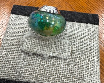 Puede incluir: Un anillo artesanal con una gran piedra de vidrio verde y azul iridiscente. La piedra está engastada en una banda de metal plateado con una parte superior decorativa. El anillo se exhibe en una pantalla de joyería de lino gris.