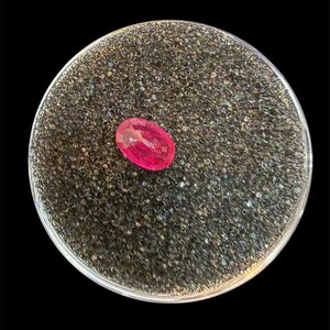 May include: A vibrant, oval-cut, hot pink gemstone rests on a circular, textured, dark gray surface. The gemstone is the focal point, showcasing its polished facets and intense color against the contrasting background. The setting is a clear, circular container.