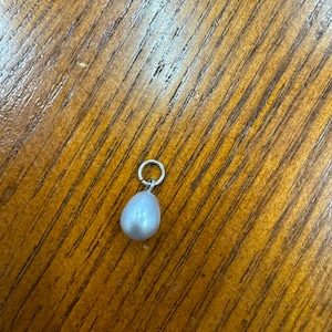 May include: A small, pear-shaped, light gray pearl pendant with a silver-colored loop. The pendant is displayed on a wooden surface with a warm, brown tone.