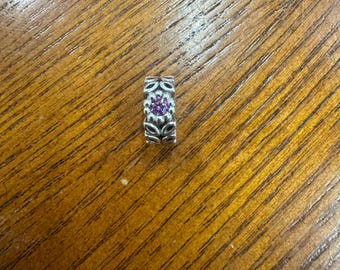 May include: A silver-colored charm with a central purple gemstone. The charm is rectangular with a floral design and leaf-like patterns. The charm is approximately 0.5 inches in length.