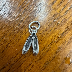 May include: Silver charm depicting two miniature ballet slippers. The charm is attached to a small ring, suitable for a bracelet or necklace. The slippers have detailed toe boxes and a polished finish, reflecting light.